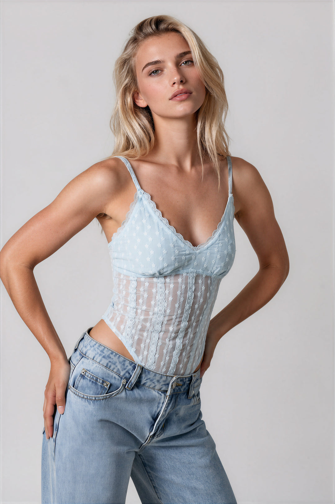 Model wearing the Gia Bodysuit in powder blue, a semi-sheer floral lace bodysuit with a scalloped V-neckline and thin adjustable straps, styled with light wash jeans