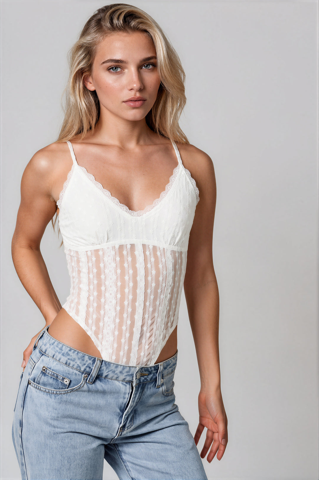 Model wearing the Gia Bodysuit in white, a semi-sheer lace bodysuit with a scalloped V-neckline and thin adjustable straps, styled with light wash jeans