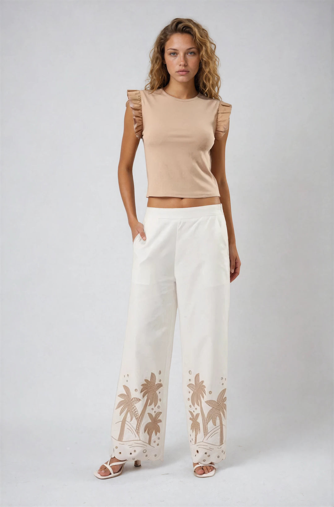 Woman wearing a beige top and white pants with palm tree designs on a plain background
