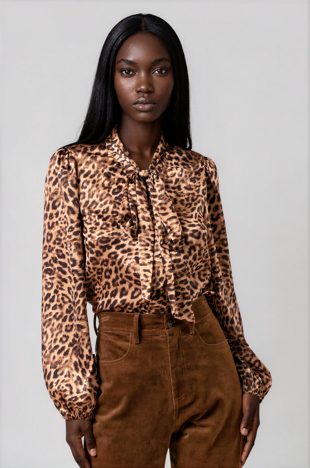 A woman modeling a long-sleeve leopard-print blouse with a ruffled tie-neck collar, paired with brown corduroy pants, against a solid light gray studio background.
