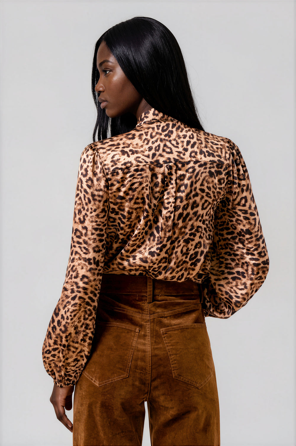 A woman shown from the back wearing a leopard-print long-sleeve blouse with a high neck and gathered shoulders, paired with brown corduroy pants, against a light gray studio background.