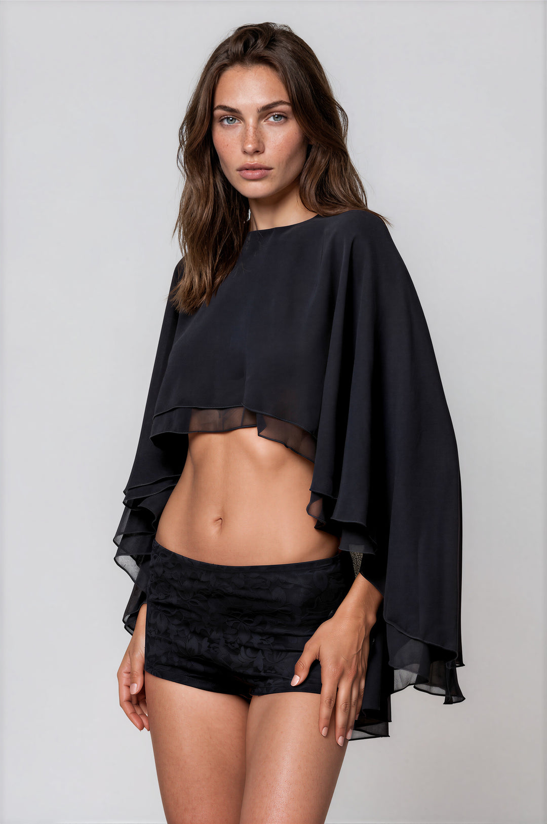 Woman wearing black cropped cape top with layered flowing sleeves and boat neckline