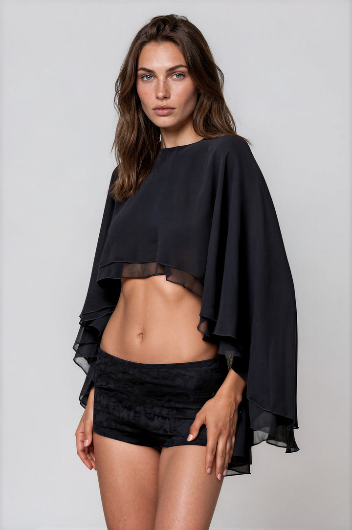 Woman wearing black cropped cape top with layered flowing sleeves and boat neckline